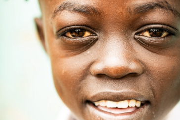 Ein Porträt eines kenianischen Kindes. Es trägt ein weißes Hemd und eine rote Kopfbedeckung, die nicht ganz zu erkennen ist. Es zeigt ein angedeutetes Lächeln.