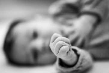 Eine schwarz-weiß-Aufnahme eines Babys. Man erkennt scharf im Vordergrund die kleine Hand, die zur Faust geballt ist. Unscharf im Hintergrund sieht man das Gesicht des Kindes.